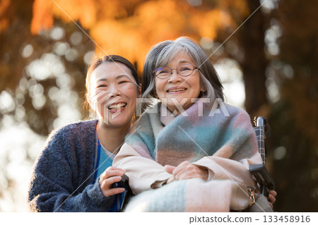一位女士正在照顧一位坐在輪椅上的老人，另一位類似護理人員的人在秋葉金黃的背景下對視，兩人都看向鏡頭。 133458916