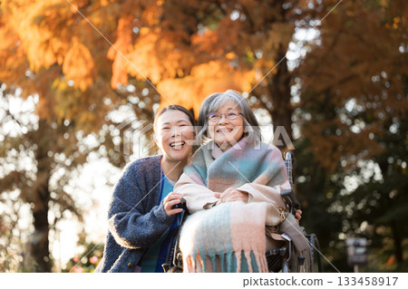 一位女士正在照顧一位坐在輪椅上的老人，另一位類似護理人員的人在秋葉金黃的背景下對視，兩人都看向鏡頭。 133458917