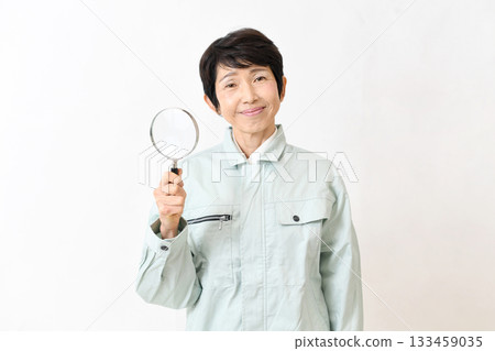 Middle-aged woman in work clothes looking at the camera with a magnifying glass Middle-aged woman in work clothes looking at the camera with a magnifying glass 133459035