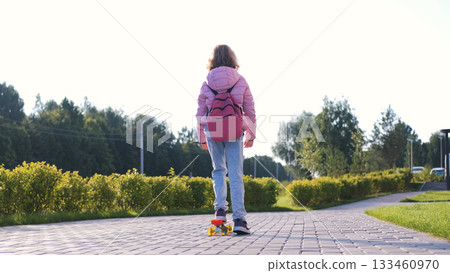 Teen girl skateboards to school in the morning wearing casual clothing and carrying a pink schoolbag gliding along a path surrounded by greenery Teen girl skateboards to school in the morning wearing casual clothing and carrying a pink schoolbag gliding along a path surrounded by greenery 133460970