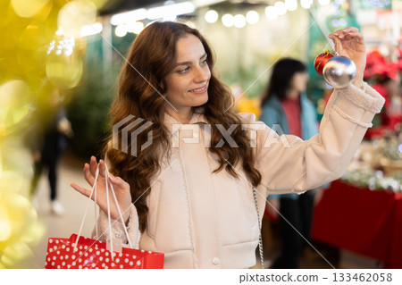 Happy woman holding Christmas baubles while walking through holiday fair Happy woman holding Christmas baubles while walking through holiday fair 133462058