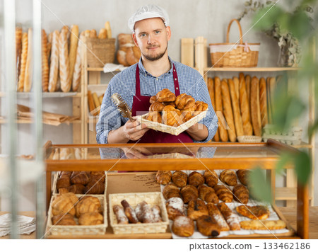 Man seller stands at bakery counter with basket filled with fresh croissants. 133462186