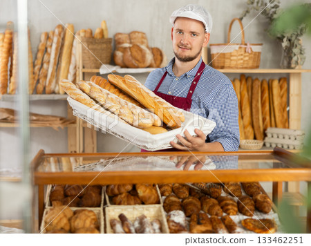 Male seller stands at bakery counter with basket filled by fresh baguettes. Male seller stands at bakery counter with basket filled by fresh baguettes. 133462251