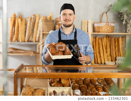Man seller stands at bakery counter with basket filled with fresh croissants. Man seller stands at bakery counter with basket filled with fresh croissants. 133462301