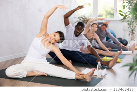 Active young woman with group doing yoga position while sitting on floor in sport club Active young woman with group doing yoga position while sitting on floor in sport club 133462455