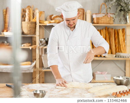 Professional baker prepares raw yeast dough in a bakery, shaping it into baguette 133462466