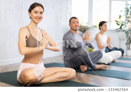 Young beautiful girl with other people relaxing and enjoying yoga in fitness center, meditation in lotus pose Young beautiful girl with other people relaxing and enjoying yoga in fitness center, meditation in lotus pose 133462564