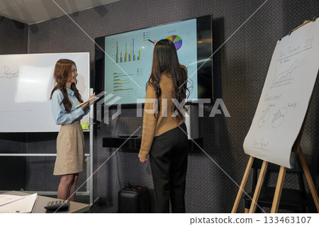 Business women meeting, busy team discuss work project, people analyze data on screen, woman points at graphs, another takes notes in modern conference room. 133463107