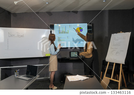 Business women meet for busy team project work during productive meeting, people collaborate in conference room, woman points at financial graphs, other takes notes for new project planning. Business women meet for busy team project work during productive meeting, people collaborate in conference room, woman points at financial graphs, other takes notes for new project planning. 133463116