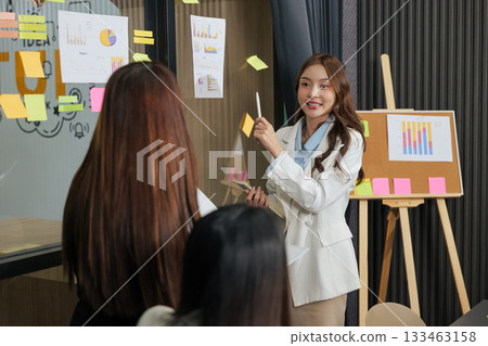 Business woman leads busy team meeting, people discuss project work, sharing ideas, presenting data visualization charts, using sticky notes for brainstorming in modern conference room. Business woman leads busy team meeting, people discuss project work, sharing ideas, presenting data visualization charts, using sticky notes for brainstorming in modern conference room. 133463158