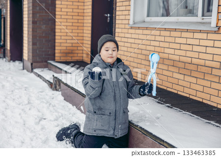 A young Asian boy kneels in the snow, holding a snowball in one hand and a snowball maker in the other. He wears a gray jacket and a beanie. A young Asian boy kneels in the snow, holding a snowball in one hand and a snowball maker in the other. He wears a gray jacket and a beanie. 133463385