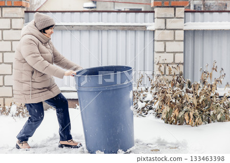 A middle-aged Asian woman in a beige coat and hat pushes a large blue trash bin through the snow in a winter setting. A middle-aged Asian woman in a beige coat and hat pushes a large blue trash bin through the snow in a winter setting. 133463398