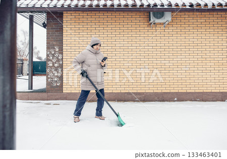 A young Asian woman in a gray winter coat shovels snow outside a brick house. He holds a smartphone in one hand while clearing the snow. A young Asian woman in a gray winter coat shovels snow outside a brick house. He holds a smartphone in one hand while clearing the snow. 133463401