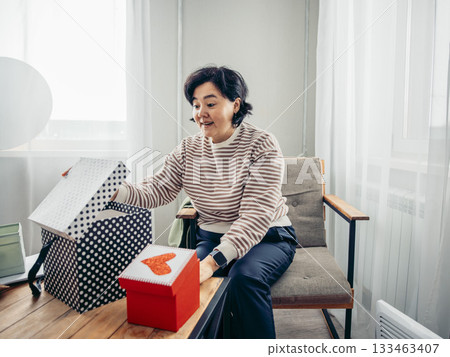 A young Asian woman with short black hair sits on a chair, opening a gift box in a cozy indoor setting. Winter Holidays: Christmas and New Year A young Asian woman with short black hair sits on a chair, opening a gift box in a cozy indoor setting. Winter Holidays: Christmas and New Year 133463407