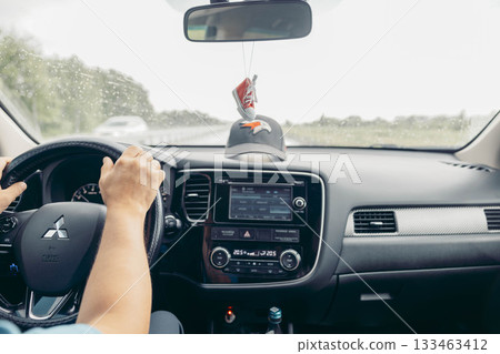 driver holds steering in car on rainy road, windshield wipers working, dashboard screen visible, front view of travel journey with mirror and hanging decoration, transport, travel, lifestyle 133463412