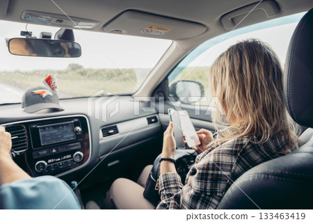 A young Caucasian woman with long blonde hair sits in a car, using her smartphone. A man drives, wearing a blue cap. The scene captures a journey on an open road. A young Caucasian woman with long blonde hair sits in a car, using her smartphone. A man drives, wearing a blue cap. The scene captures a journey on an open road. 133463419