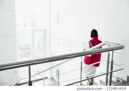 Unrecognizable woman in a red blazer walks down a modern staircase in a bright office environment. She carries a laptop and appears to be on a break. Unrecognizable woman in a red blazer walks down a modern staircase in a bright office environment. She carries a laptop and appears to be on a break. 133463454