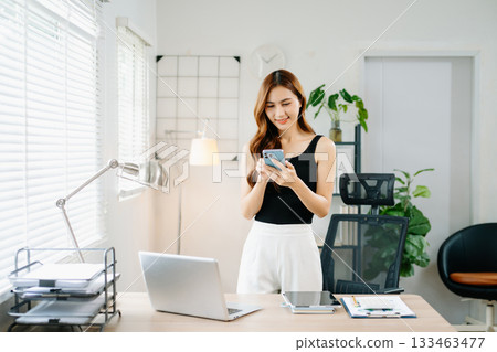 Smiling Asian woman standing in bright modern home office using smartphone, concept of digital lifestyle, communication, and remote working success. Smiling Asian woman standing in bright modern home office using smartphone, concept of digital lifestyle, communication, and remote working success. 133463477