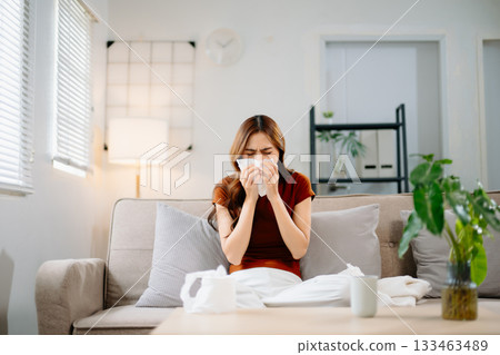 Young Asian woman suffering from flu symptoms, covered with a blanket on a sofa. Concept of illness, healthcare, and recovery. Stay indoors, stay healthy 133463489