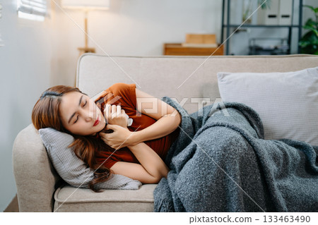 Young Asian woman suffering from flu symptoms, covered with a blanket on a sofa. Concept of illness, healthcare, and recovery. Stay indoors, stay healthy 133463490