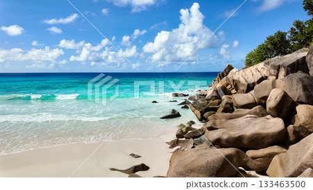 Granite boulders on a white sandy beach with turquoise waves washing ashore under a bright blue sky. Seychelles, La Digue. Grand Anse. 133463500