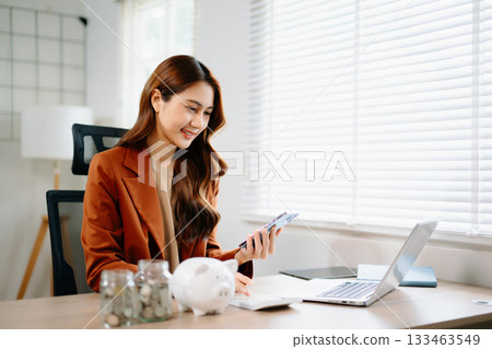 Asian businesswoman using smartphone and calculator for finance planning with piggy bank, coin jar, and laptop on desk 133463549