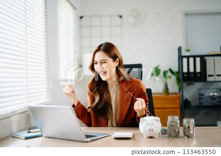 Happy Asian businesswoman celebrating financial success at office desk with laptop, piggy bank, and coin jars, symbolizing savings, investment, fintech, and career growth. Happy Asian businesswoman celebrating financial success at office desk with laptop, piggy bank, and coin jars, symbolizing savings, investment, fintech, and career growth. 133463550