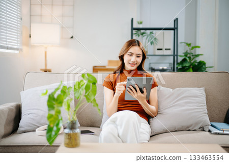 Happy Asian woman sitting on sofa and using digital tablet at home. Modern lifestyle, online learning, and technology concept for remote work and digital connection. 133463554