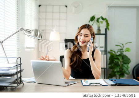 Smiling businesswoman talking on phone while working on laptop in bright modern office, symbolizing success, communication 133463591
