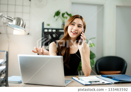 Smiling businesswoman talking on phone while working on laptop in bright modern office, symbolizing success, communication Smiling businesswoman talking on phone while working on laptop in bright modern office, symbolizing success, communication 133463592