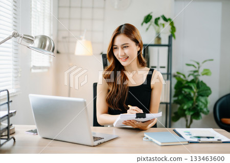 Smiling Asian woman standing and working with laptop in modern home office, concept of productivity, success, and female entrepreneurship. 133463600
