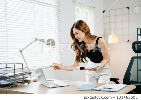 Smiling Asian woman standing and working with laptop in modern home office, concept of productivity, success, and female entrepreneurship. 133463603