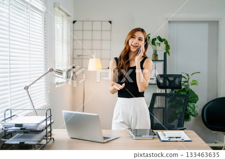 Smiling businesswoman talking on phone while working on laptop in bright modern office, symbolizing success, communication Smiling businesswoman talking on phone while working on laptop in bright modern office, symbolizing success, communication 133463635