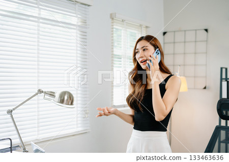 Smiling businesswoman talking on phone while working on laptop in bright modern office, symbolizing success, communication Smiling businesswoman talking on phone while working on laptop in bright modern office, symbolizing success, communication 133463636