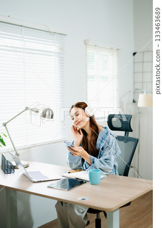 Cheerful woman wearing headphones using smartphone beside laptop, symbolizing digital lifestyle, communication, creativity Cheerful woman wearing headphones using smartphone beside laptop, symbolizing digital lifestyle, communication, creativity 133463689