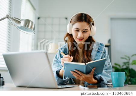 Smiling woman studying online with laptop and headphones at home, holding notebook in bright workspace, symbolizing education and digital lifestyle. 133463694
