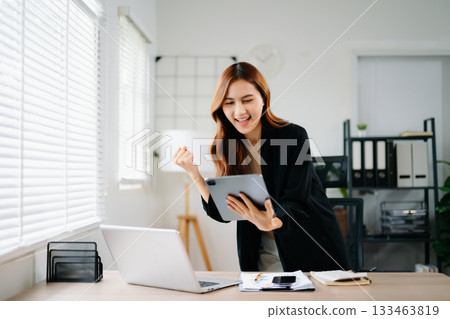 Asian businesswoman celebrating success at work, standing and cheering in modern office with laptop, showing joy, achievement, and victory in business and digital career. Asian businesswoman celebrating success at work, standing and cheering in modern office with laptop, showing joy, achievement, and victory in business and digital career. 133463819