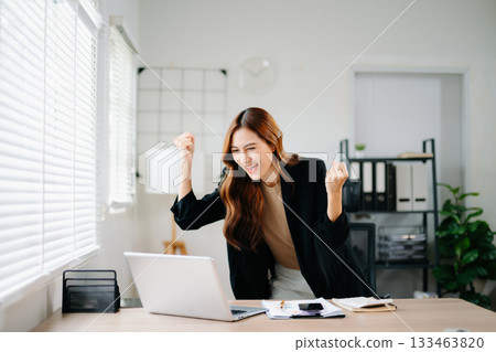 Asian businesswoman celebrating success at work, standing and cheering in modern office with laptop, showing joy, achievement, and victory in business and digital career. 133463820