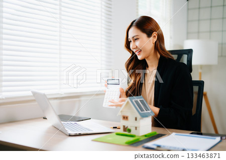 Young real estate agent worker working with laptop and tablet at table in office and small house beside it. Young real estate agent worker working with laptop and tablet at table in office and small house beside it. 133463871
