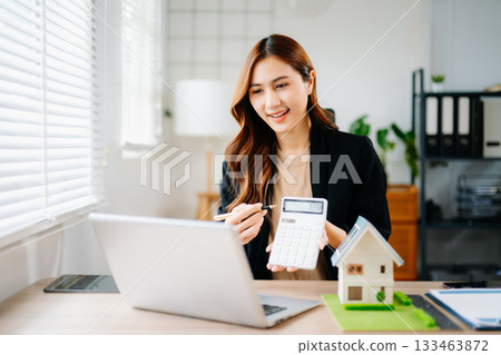 Asian real estate agent using smartphone in modern office with house model and laptop, representing property sales, investment, mortgage consulting, and housing business. 133463872