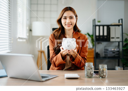 Smiling Asian businesswoman holding piggy bank at office desk with coin jars and laptop, symbolizing savings, smart money, financial planning 133463927