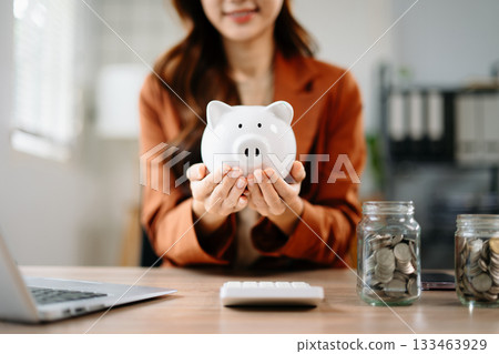 Smiling Asian businesswoman holding piggy bank at office desk with coin jars and laptop, symbolizing savings, smart money, financial planning 133463929