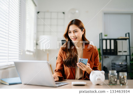 Smiling businesswoman saves money with piggy bank and coins. Ideal for finance, savings, budgeting, and office Smiling businesswoman saves money with piggy bank and coins. Ideal for finance, savings, budgeting, and office 133463934