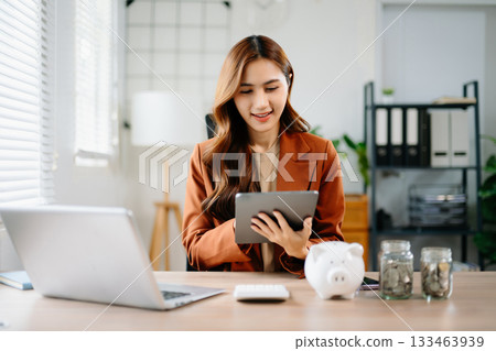 Young Asian businesswoman using tablet for finance and savings planning in modern office with piggy bank and coin jars, symbolizing money management, fintech, and success. Young Asian businesswoman using tablet for finance and savings planning in modern office with piggy bank and coin jars, symbolizing money management, fintech, and success. 133463939