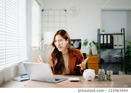 Happy Asian businesswoman celebrating financial success at office desk with laptop, piggy bank, and coin jars, symbolizing savings, investment, fintech, and career growth. Happy Asian businesswoman celebrating financial success at office desk with laptop, piggy bank, and coin jars, symbolizing savings, investment, fintech, and career growth. 133463944