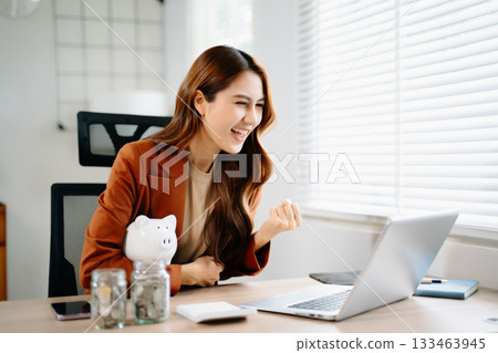 Happy Asian businesswoman celebrating financial success at office desk with laptop, piggy bank, and coin jars, symbolizing savings, investment, fintech, and career growth. 133463945