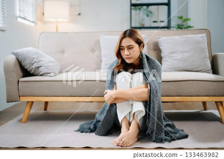 Sad Asian woman sitting on floor wrapped in blanket, holding head in distress. Concept of stress, anxiety, depression, burnout, and mental health problems. 133463982