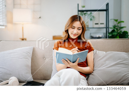 Smiling Asian woman sitting on sofa, thinking and writing notes in notebook. Creative idea, planning, education, and inspiration concept in modern home environment. Smiling Asian woman sitting on sofa, thinking and writing notes in notebook. Creative idea, planning, education, and inspiration concept in modern home environment. 133464059