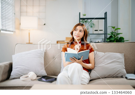 Smiling Asian woman sitting on sofa, thinking and writing notes in notebook. Creative idea, planning, education, and inspiration concept in modern home environment. Smiling Asian woman sitting on sofa, thinking and writing notes in notebook. Creative idea, planning, education, and inspiration concept in modern home environment. 133464060