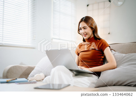 Cheerful Asian woman smiling while working on laptop at home. Modern remote work, online learning, and freelance lifestyle concept with confidence and creativity. 133464062